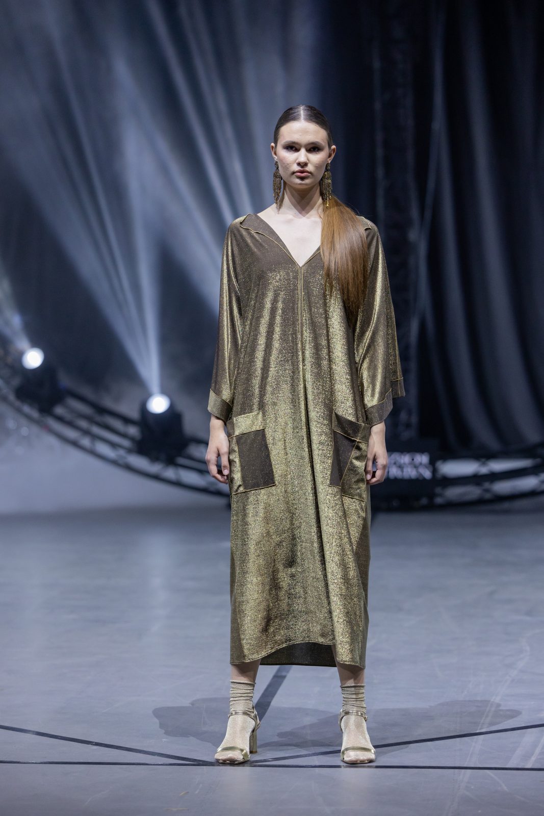 Модель в длинном золотом платье с карманами на фоне сценических огней на неделе моды в Беларуси.