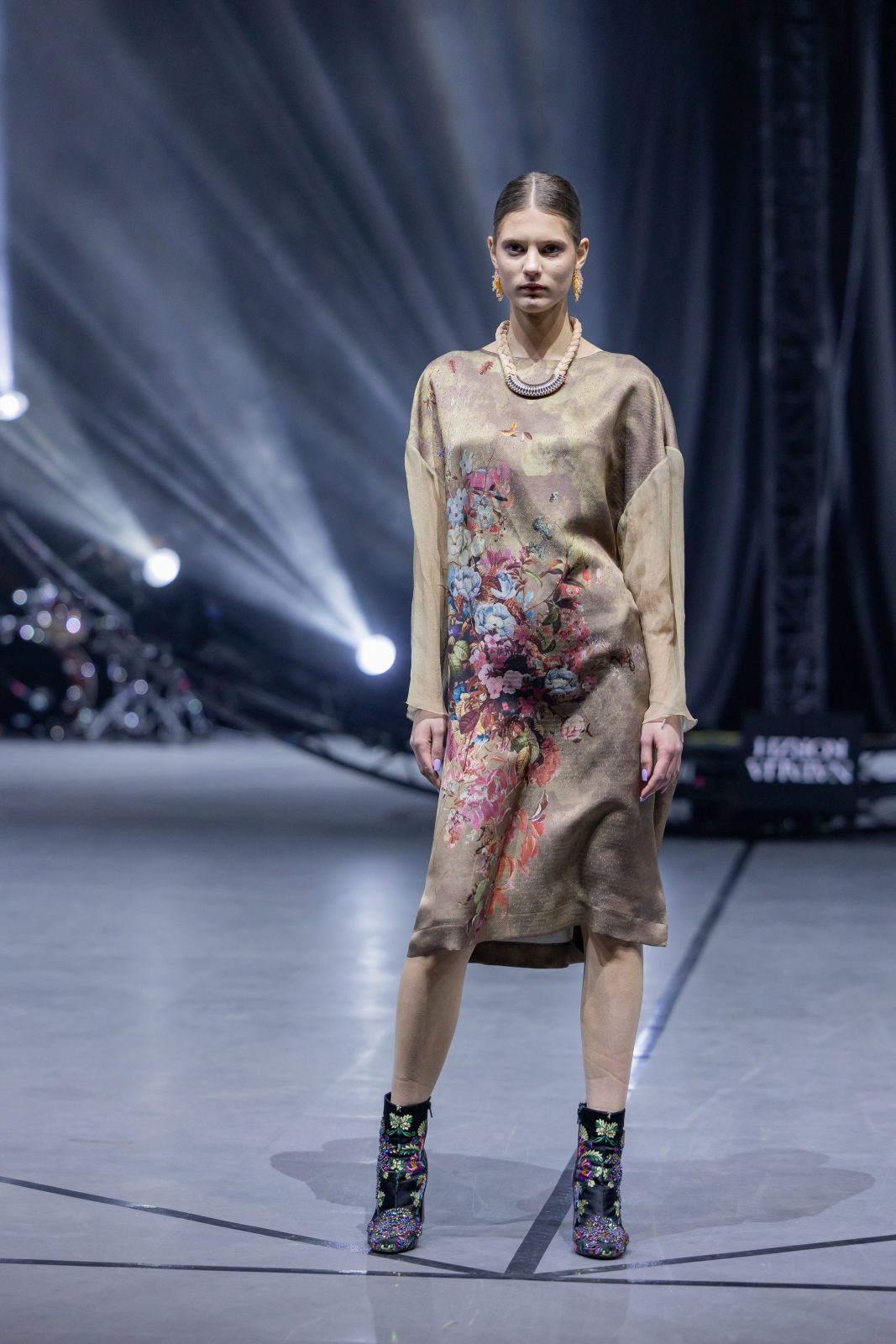 Модель на подиуме во время Belarus Fashion Art Week, одета в длинное платье с цветочным принтом и стильными обувью. На фоне световые эффекты.