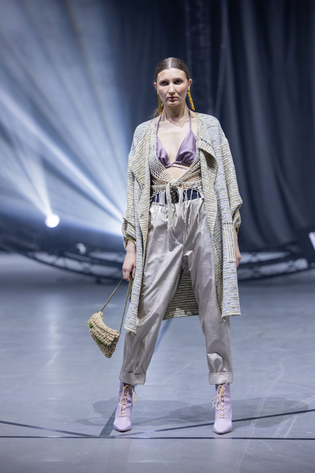 Модель на подиуме во время Belarus Fashion Art Week, одета в стильный наряд с золотистыми брюками, облегающим топом и кардиганом, держа в руках небольшую сумку.