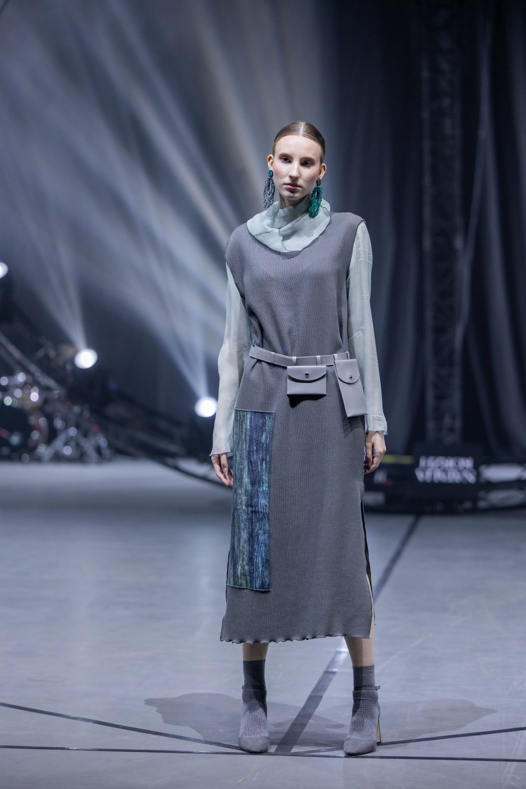 Модель на подиуме во время Belarus Fashion Art Week, одетая в современное серое платье с яркими акцентами и декоративными элементами.
